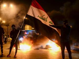 Masked anti-government protester wave the national flag before flaming tyres at a make-shift roadblock in the central Iraqi holy shrine city of Najaf on February 1, 2020. Iraq's president named former communications minister Mohammad Allawi as the country's new prime minister after an 11th-hour consensus among political blocs, but the streets seemed divided on his nomination. Haidar HAMDANI / AFP
