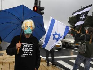 A man holding an umbrella, wearing a protective face mask beneath another mask showing the face of late physicist Albert Einstein, and a hoodie sweater quoting a verse from the biblical book of Isaiah, while another waves an Israeli flag and another Israeli flag with the colours inverted during a protest outside the Knesset (parliament) in Jerusalem on March 19, 2020. AFP/File