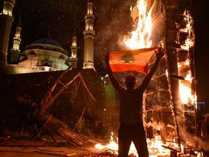 A Lebanese man raises a national flag as the ‘Revolution fist’, symbol of Lebanon’s October 2019 uprising, burns after being torched during clashes between anti-government protesters and supporters of former prime minister Saad Hariri, in the capital Beirut's central Martyr's square, on October 21, 2020. Hariri resigned as premier in October 2019 in the wake of unprecedented street protests, but he is now expected to make a comeback at the helm of the next government. Most parliamentary blocs have pledged s
