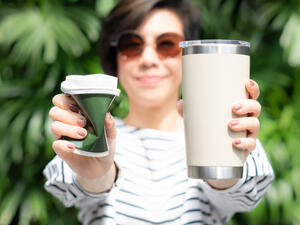 Woman holds takeaway coffee cup in both hands, one is a single use paper cup with plastic lid, the other one is a reusable stainless tumbler