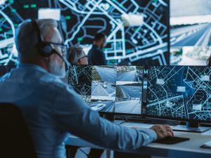 Technical Customer Support Specialist is Talking on a Headset while Working on a Computer in a Call Center Control Room Filled Display Screens with Monitoring Software. Over the Shoulder Footage