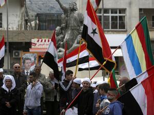 Residents of the Golan Heights wave Syrian and Druze flags as they gather in front of a portrait of the Syrian President Bashar al-Assad during a protest against the backing of Israel's capture of the Golan Heights by the US president, in the village of Majdal Shams in the Israeli-annexed territory (AFP)
