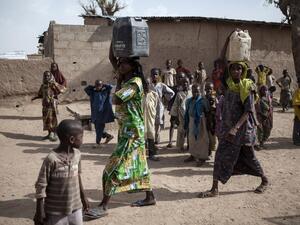 Two women carrying jerrycans filled with water walk past children in the town of Banki in northeastern Nigeria on April 26, 2017. (AFP/Florian Plaucheur)