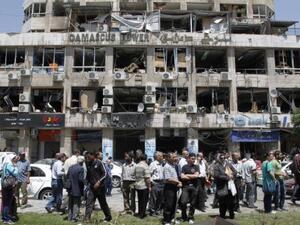 People stand on a street lined with a damaged building and destroyed cars after a blast at Marjeh Square in Damascus in April. Image courtesy of nydailynews.com
