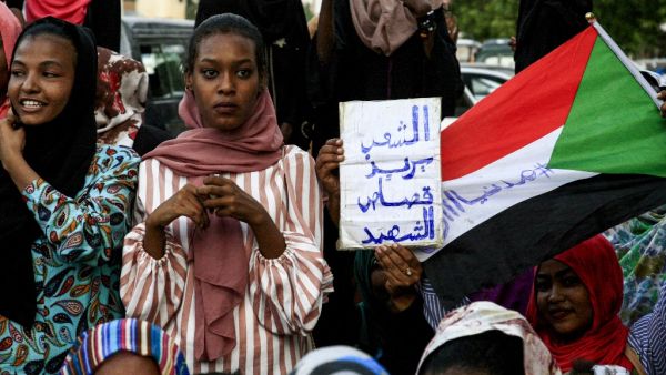 A Sudanese woman holds up a sign reading in Arabic "the people want retribution for the martyrs" next to a waving national flag defaced with the Arabic text "#CiviliaNow". (AFP/ File Photo)