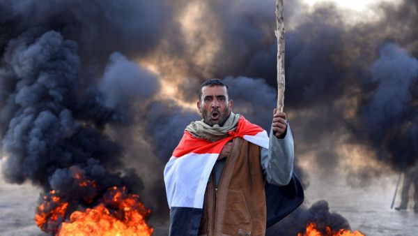 An anti-government protester draped in a national flag chants slogans while holding a stick at a make-shift roadblock with flaming tyres during a demonstration against the new Iraqi prime minister designate in the central holy shrine city of Najaf on February 2, 2020. AFP