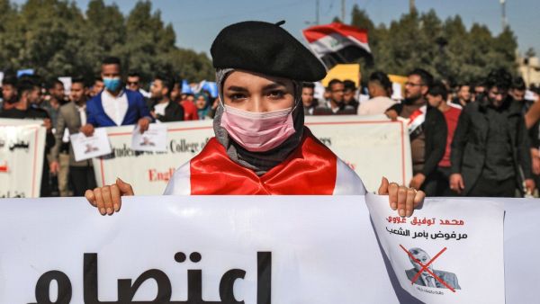 In this file photo taken on February 2, 2020, An Iraqi student wearing a beret and a surgical mask holds signs with a red X mark on the face of the new Iraqi prime minister designate Mohammad Allawi reading in Arabic "rejected by order of the people" during a demonstration against him in the central holy shrine city of Karbala. (AFP)
