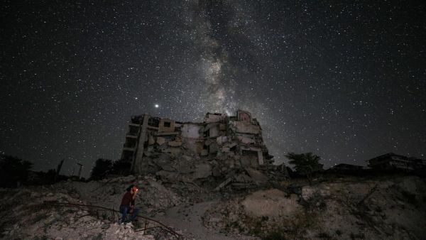This long-exposure picture taken early on June 27, 2020 shows a man smoking past buildings destroyed by prior bombardment in the town of Ariha in Syria's rebel-held northwestern Idlib province, as the Milky Way galaxy is seen in the night sky above. Omar HAJ KADOUR / AFP