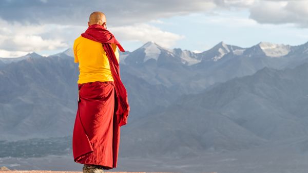 Monk lama in red and yellow color clothing standing in front of mountains (Shutterstock)	 