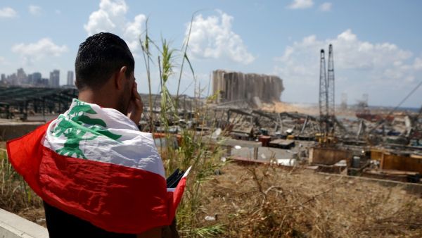 A man draped in a Lebanese flag reacts as he stands before the ravaged port of Lebanon's capital Beirut on August 9, 2020, in the aftermath of a colossal explosion that occurred days prior due to a huge pile of ammonium nitrate that had languished for years at a port warehouse. The huge chemical explosion that hit Beirut's port, devastating large parts of the Lebanese capital and claiming over 150 lives, left a 43-metre (141 foot) deep crater, a security official said. The blast Tuesday, which was felt acro