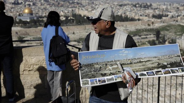 A vendor sells souvenir postcards to tourists watching Jerusalem's Old City from the panoramic view of the Mount of Olives, January 2019 (AFP) A vendor sells souvenir postcards to tourists watching Jerusalem's Old City from the panoramic view of the Mount of Olives, January 2019 (AFP)