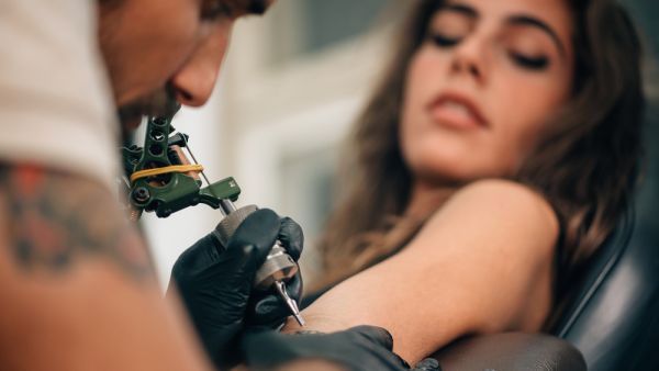 Tattoo artist creating a tattoo on a girl's arm  (Shutterstock)	 What is the Tattoo artist doing?