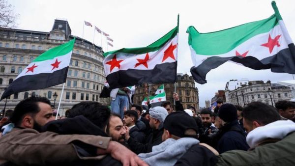 متظاهرون يرفعون أعلام الثورة السورية في ساحة عامة وسط تجمع حاشد، مع مبانٍ تاريخية في الخلفية.