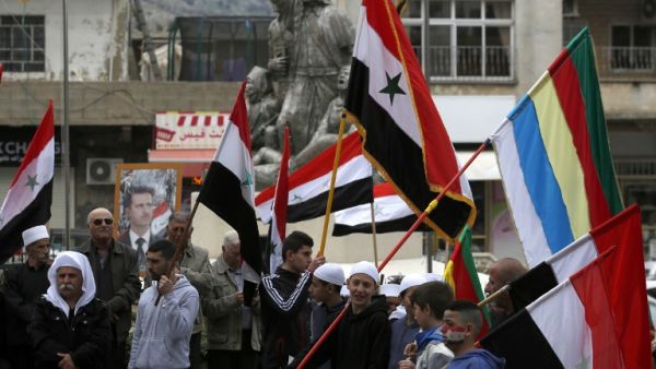 Residents of the Golan Heights wave Syrian and Druze flags as they gather in front of a portrait of the Syrian President Bashar al-Assad during a protest against the backing of Israel's capture of the Golan Heights by the US president, in the village of Majdal Shams in the Israeli-annexed territory (AFP)