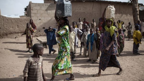 Two women carrying jerrycans filled with water walk past children in the town of Banki in northeastern Nigeria on April 26, 2017. (AFP/Florian Plaucheur)