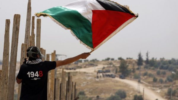 A Palestinian protester, wearing a gas mask, waves a Palestinian flag in front of Israeli security forces on May 13, 2016 in the West Bank village of Bilin, near Ramallah, during a march against the building of Israel's controversial separation barrier and the construction of Israel settlements. (AFP/Abbas Momani)