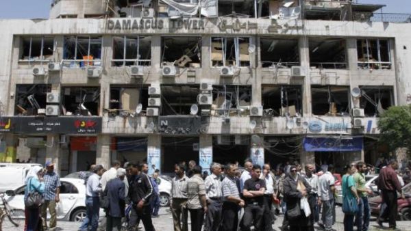 People stand on a street lined with a damaged building and destroyed cars after a blast at Marjeh Square in Damascus in April. Image courtesy of nydailynews.com
