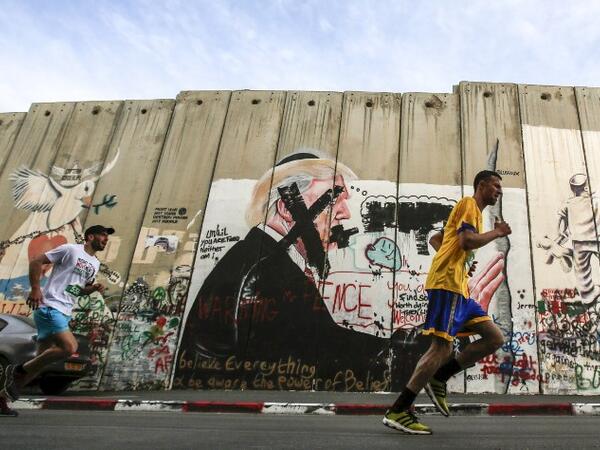 Participants run past a mural depicting U.S. President Donald Trump drawn along Israel's controversial separation barrier, which divides the West Bank from Jerusalem, in the biblical town of Bethlehem during the 6th International Palestine Marathon on Mar. 23, 2018. 
(Musa AL SHAER / AFP)