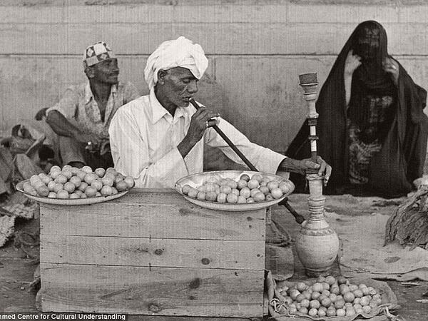 A man selling dried herbs and lemon pauses to smoke a shisha pipe, ca. 1960s
