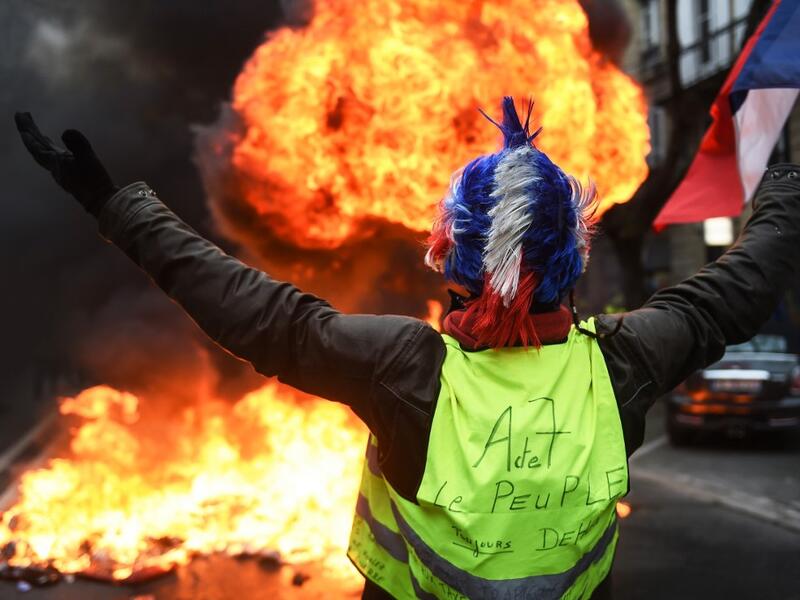 In this file photo taken on December 29, 2018 A "Yellow vest" (gilets jaunes) anti-government demonstrator holds up the French flag as he stands infront of a fire during protests in the western French city of Bordeaux. On November 17, 2018, 282,000 people according to the authorities, responded to a Facebook call, yellow vest on their backs, outside any political or trade union framework, and invested hundreds of roundabouts, symbols of the peri-urban France. The movement, born after a contested planned fue