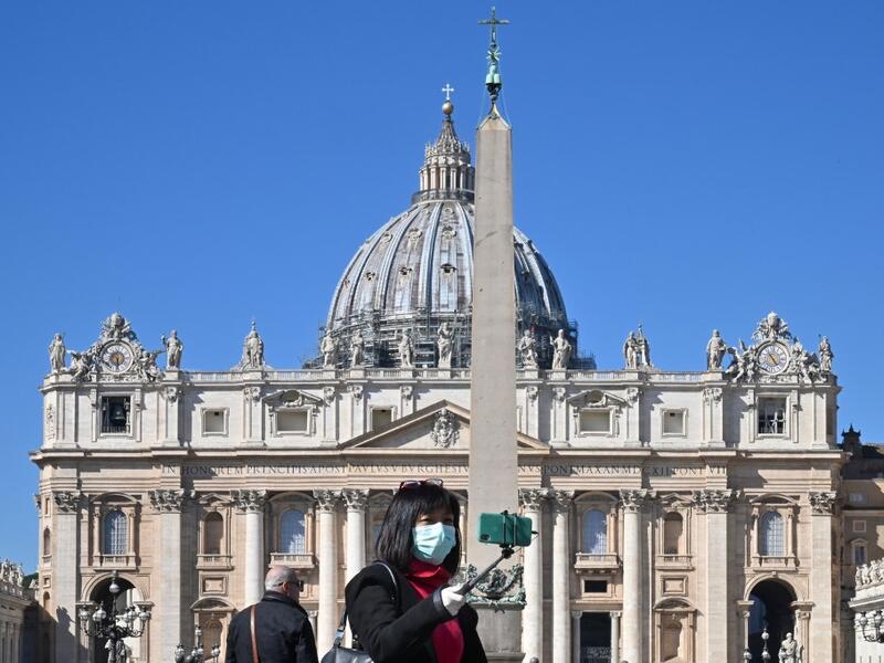 A photo taken on March 11, 2020 shows a tourist wearing a protective mask taking a selfie photo by the Vatican's Saint Peter's Square and its main basilica, a day after they were closed to tourists as part of a broader clampdown aimed at curbing the coronavirus outbreak. ANDREAS SOLARO / AFP