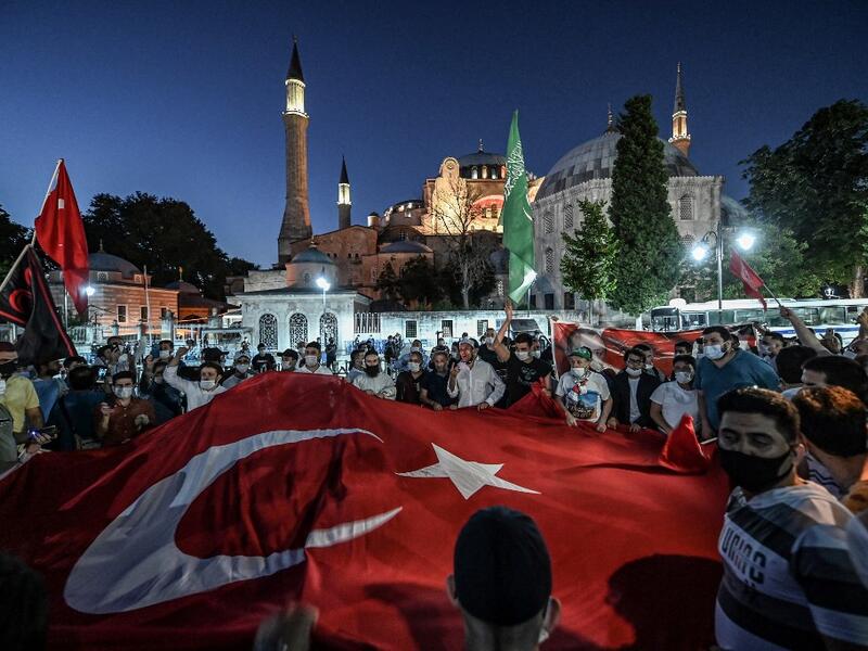 People, some wearing face masks, wave a giant Turkey national flag and shout slogans outside the Hagia Sophia museum in Istanbul on July 10, 2020 as they gather to celebrate after a top Turkish court revoked the sixth-century Hagia Sophia's status as a museum, clearing the way for it to be turned back into a mosque. The Council of State, the country's highest administrative court which on July 2 debated a case brought by a Turkish NGO, cancelled a 1934 cabinet decision and ruled the UNESCO World Heritage si