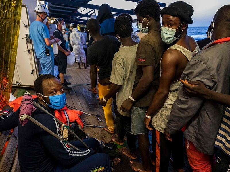 Migrants who have just been helped off the Louise Michel queue to see crew members and doctors including an injured man with crutches (AFP)