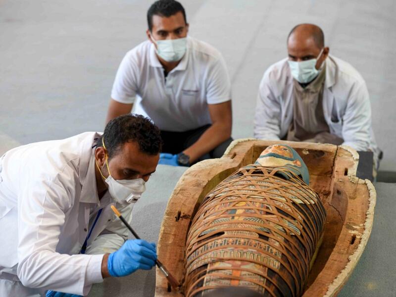 Archaeologists inspect a mummy, wrapped in a burial shroud adorned with brightly coloured hieroglyphic pictorials, during the unveiling of an ancient treasure trove of more than a 100 intact sarcophagi, at the Saqqara necropolis 30 kms south of the Egyptian capital Cairo, on November 14, 2020. Egypt announced the discovery of an ancient treasure trove of more than a 100 intact sarcophagi, the largest such find this year. The sealed wooden coffins, unveiled on site amid fanfare, belonged to top officials of 