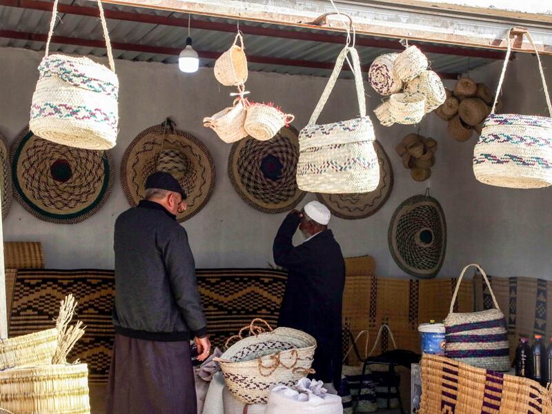 People inspect palm wicker products on display, made by Haleema Mohamed, one of the residents who returned to the city of Tawergha after fleeing in the aftermath of the toppling of Kadhafi, sits making palm wicker products at a home in Tawergha, some 200 kilometres (125 miles) east of Libya's capital close to the port city of Misrata, on December 12, 2020. When Libyan dictator Moamer Kadhafi was toppled, people took revenge on those they saw as his supporters -- including the entire town of Tawergha, whose 