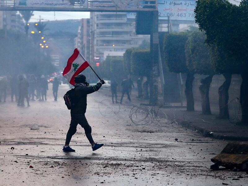 A Lebanese anti-government protester waves a national flag during clashes with security forces in front of the Serail (headquarters of the Governorate), in the northern port city of Tripoli, following a demonstration to protest against the economic situation, on January 28, 2021. Tripoli was already one of Lebanon's poorest areas before the coronavirus pandemic piled new misery onto a chronic economic crisis. Many of its residents have been left without an income since Lebanon imposed a full lockdown earlie