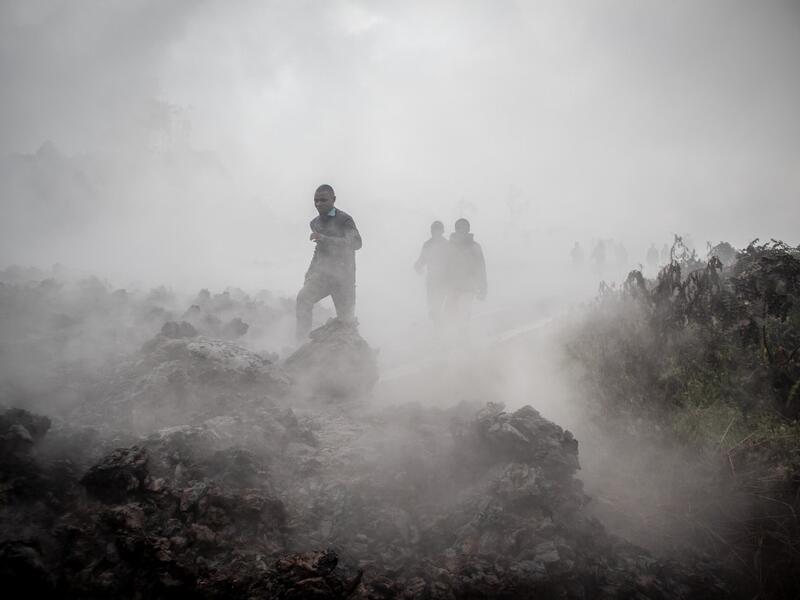 Men cross the front of the still smoking lava rocks from an eruption of the Mount Nyiragongo on May 23, 2021 in Goma in the East of the Democratic Republic of Congo. A river of boiling lava came to a halt on the outskirts of Goma Sunday, sparing the city in eastern DR Congo from disaster after the nighttime eruption of Mount Nyiragongo sent thousands of terrified residents fleeing in panic. Fire and strong fumes emanated from the blackish molten rock as it swallowed up houses
