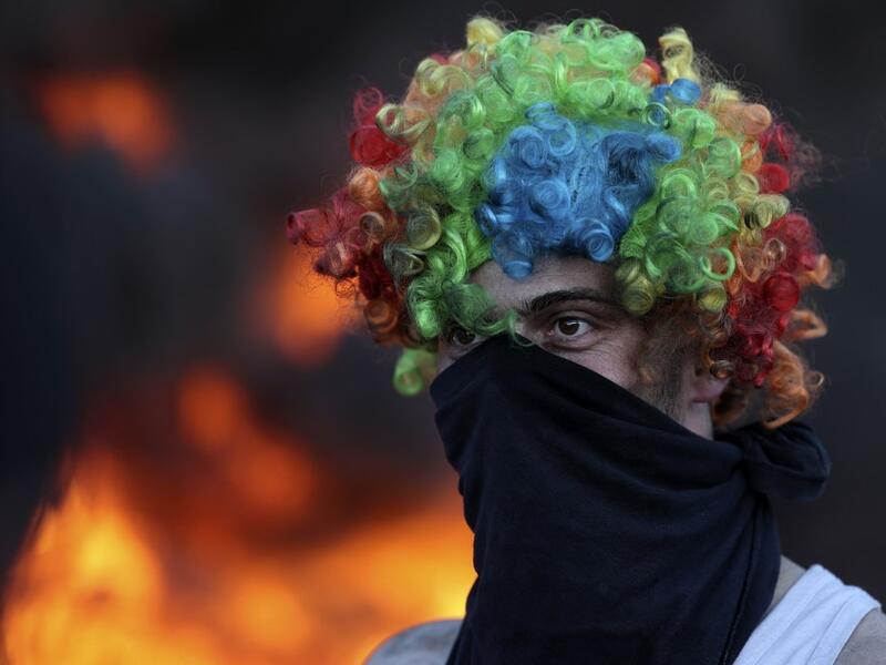 A man wears a face cover and a colorful wig as Palestinians burn tires during a night demonstration the expansion of the Jewish settlement outpost of Eviatar on the lands of Beita village, near the occupied West Bank city of Nablus