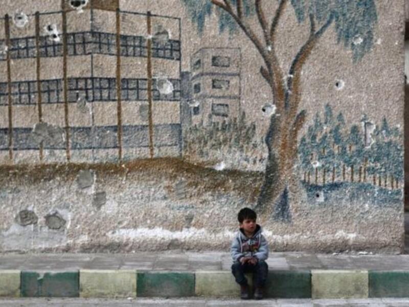 A Syrian child sits in front of a mural covered in bullet holes on the wall. (AFP/File)