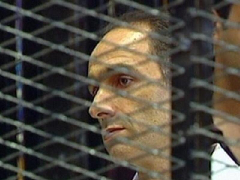 Gamal Mubarak is seen in a holding cell on the first day of his trial along with his brother and father, ousted Egyptian president Mubarak and other government officials. Father-on-trial said to be fidgety & squirming, even playing with his nose; some have irreverently cited him as 'picking' it.