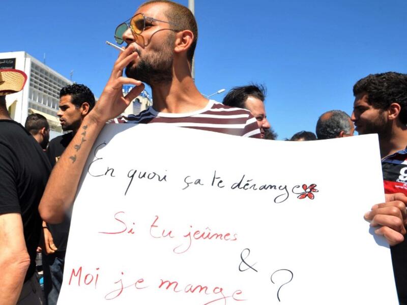 A Tunisian protester smokes a cigarette and holds a placard reading in French "Why is it bothering you? If you fast and I eat?" during a demonstration for the right to eat and smoke in public during the Muslim dawn-to-dusk fasting month of Ramadan, on June 11, 2017, in Tunis. (Sofienne Hamdaoui/AFP)