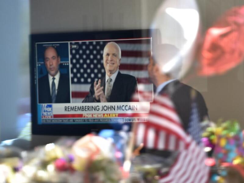 A man in the lobby of Senator John McCain's former office building watches a Fox News report as flowers and balloons from a makeshift memorial located outside the building are reflected in the building's glass window. (Robyn Beck / AFP)