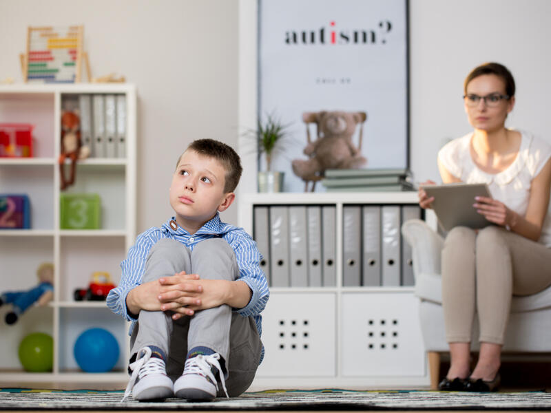 Psychologist in glasses is looking at kid sick of autism sitting on carpet in classroom. (Shutterstock/ File Photo)