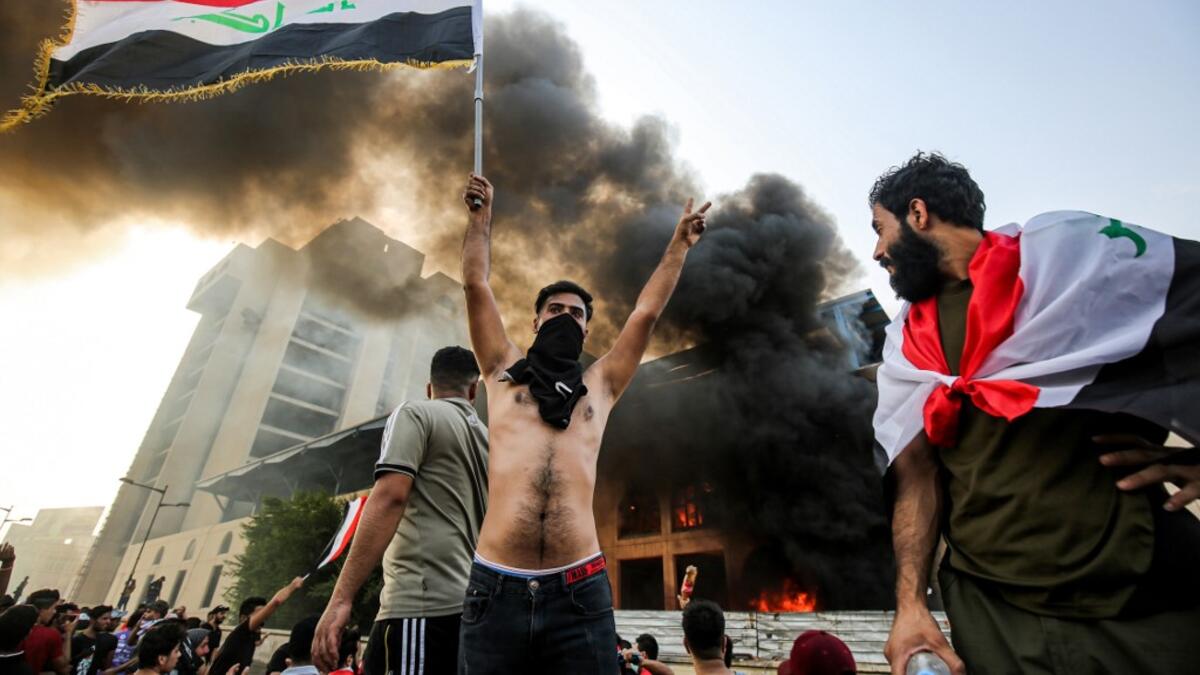 A topless masked protester flashes the victory gesture as he waves an Iraqi national flag before a burning building during a demonstration against state corruption and poor services, between the capital Baghdad's Tahrir Square and the high-security Green Zone district, on October 1, 2019.  AHMAD AL-RUBAYE / AFP