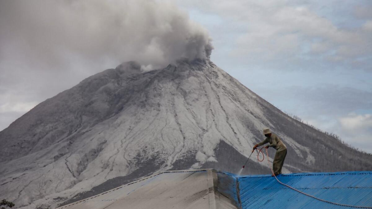 A villager cleans volcanic ash from his roof at Naman Teran village in Karo, North Sumatra, on August 11, 2020 a day after the eruption of Mount Sinabung (background). IVAN DAMANIK / AFP