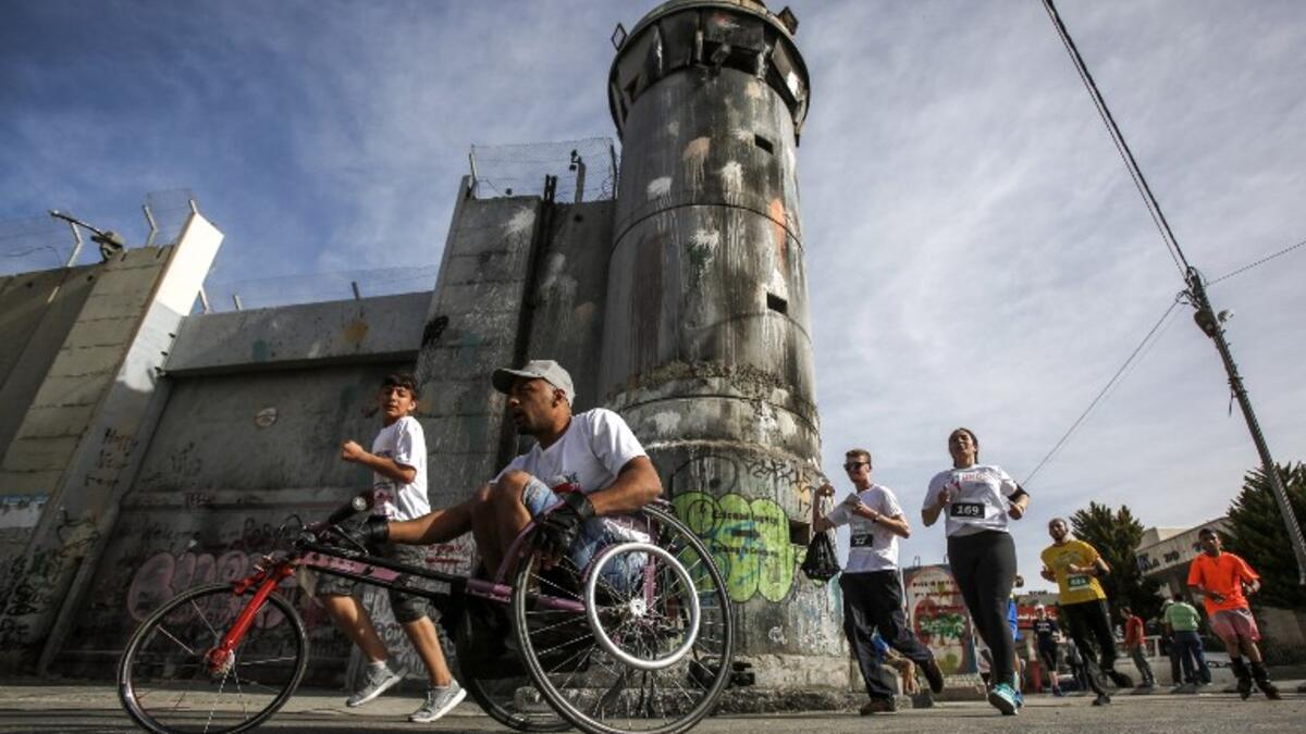A participant rides in a wheelchair with others running along Israel's controversial separation barrier, which divides the West Bank from Jerusalem, in the biblical town of Bethlehem during the 6th International Palestine Marathon on Mar. 23, 2018. 
(Musa AL SHAER / AFP)