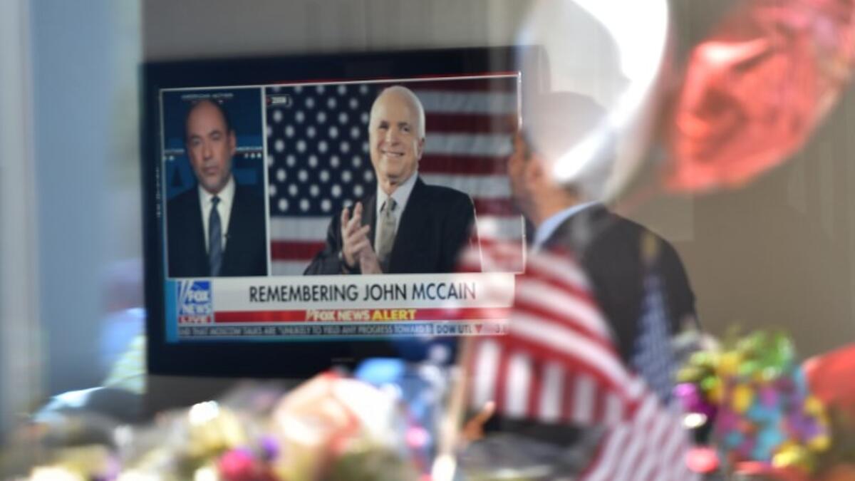 A man in the lobby of Senator John McCain's former office building watches a Fox News report as flowers and balloons from a makeshift memorial located outside the building are reflected in the building's glass window. (Robyn Beck / AFP)